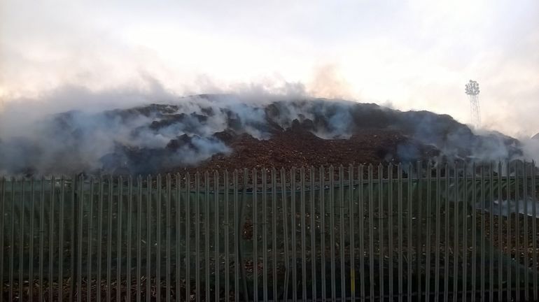 Smoke bellowing from a pile of wood chip surrounded by a metal fence