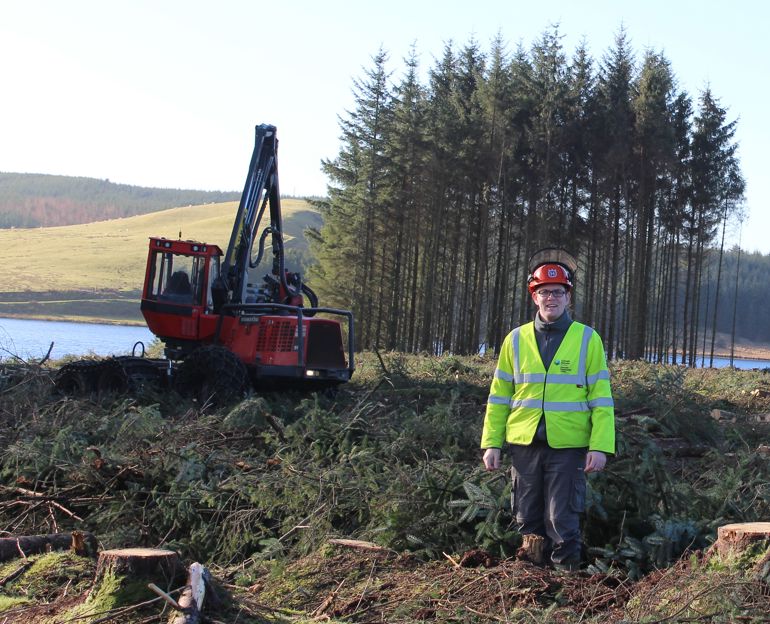 Dyn mewn siaced hawdd-ei-gweld yn sefyll o flaen peiriant cloddio a chlwstwr o goed