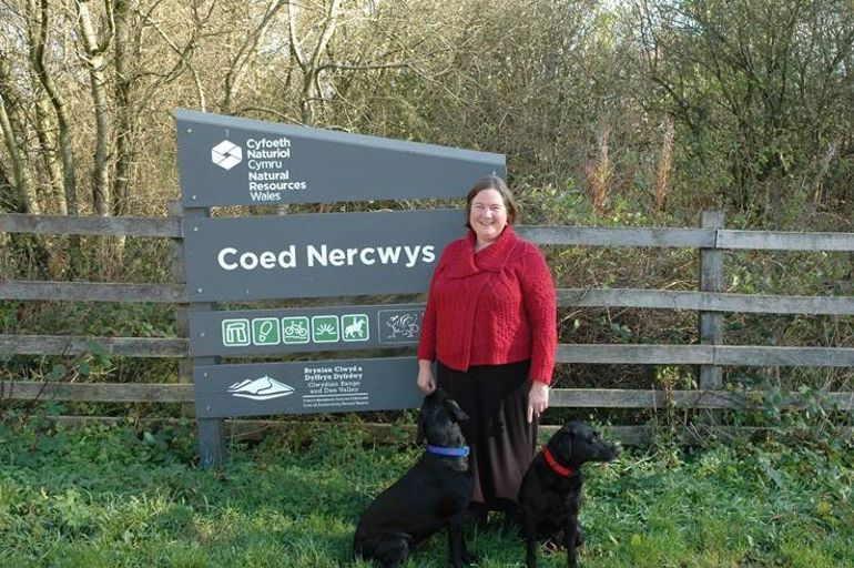 Clare Pillman CEO, wearing red jacket stood in front of the NRW Coed Nercwys sign with two black labrador dogs