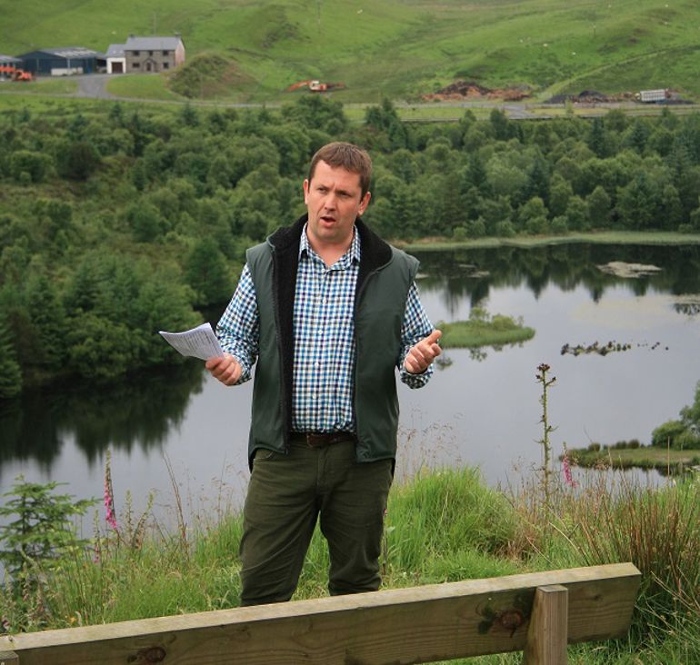 Man in checked shirt and waistcoat stood at the side of a lake with piece of paper in his hand