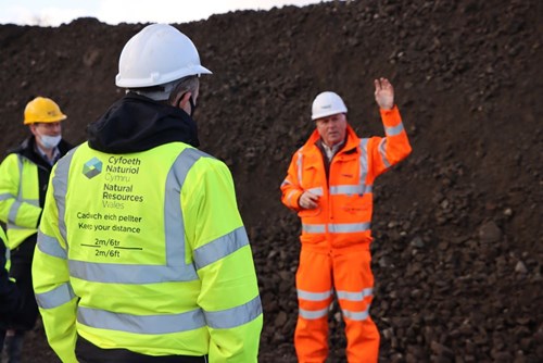 Image of three workers. The focus is on the person in the foregrounds jacket which has 'Keep your distance' on the back